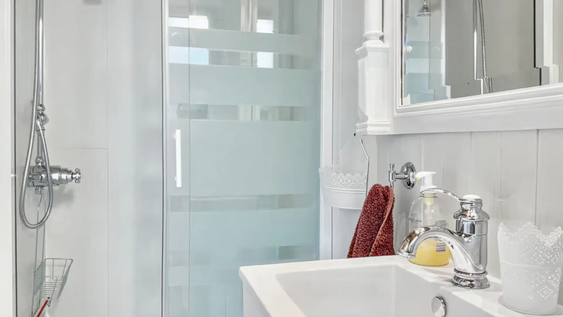 Un gros plan d'une salle de bain moderne et lumineuse, décorée dans des tons de blanc. Au premier plan et à droite, un lavabo carré en céramique blanche est intégré dans un meuble dont la surface est en marbre blanc. Le robinet est chromé et brillant. À côté du lavabo, une bouteille de savon liquide et un porte-serviettes en éponge couleur rouille sont visibles. Le mur derrière le lavabo est recouvert de lambris blanc. À gauche, une cabine de douche est fermée par une paroi de verre avec des bandes horizontales dépolies. À l'intérieur de la douche, une pomme de douche chromée est fixée au mur. Le meuble-miroir au-dessus du lavabo est également blanc et possède des moulures décoratives.