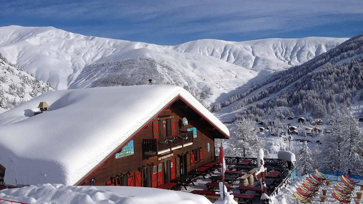 View from the restaurant, a wooden chalet with a terrace, tables and chairs, and long chairs. Snow-capped mountains all around and in the background.