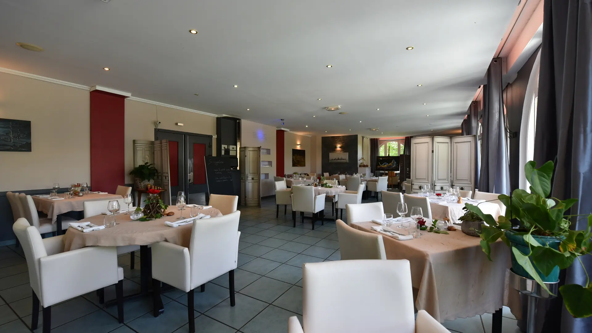 Une dizaine de tables sont dressées pour le repas dans une salle de restauration spacieuse décorée avec des tableaux et des plantes vertes