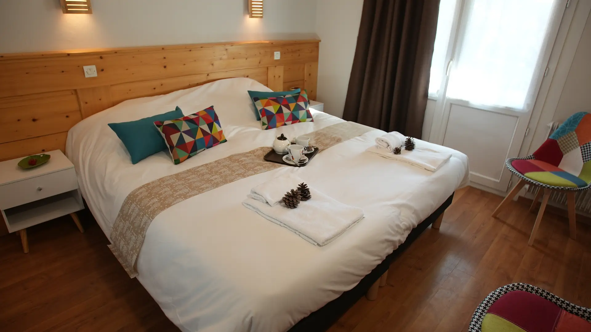 Lit double avec coussins décoratifs et serviettes blanches, un plateau y est posé avec tasses et théière. Deux chevets encadrent le lit et deux sièges patchwork y font face