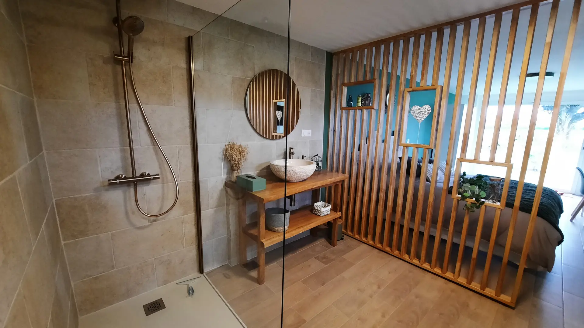 Vue depuis la salle d'eau où se trouve une large douche à l'italienne. Un espace lumineux prévu pour la douche est séparé de la chambre par une haute cloison en bois. Un lavabo, des rangements et un miroir complètent le coin de la pièce.