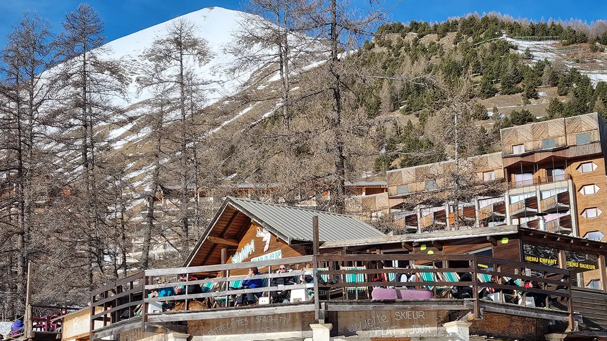 Vue d'ensemble du restaurant en bois, situé devant des immeubles de la station de ski, en haut d'un bâtiment sur deux niveaux, terrasse. Panneau Sympa Snack sur la devanture. Montagnes en arrière-plan