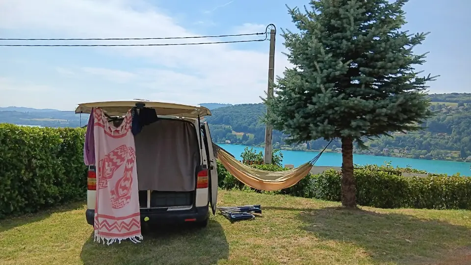 Photo d'un camping car parké sur un emplacement de camping herboré, avec un hamac et une jolie vue dégagée sur le lac de paladru