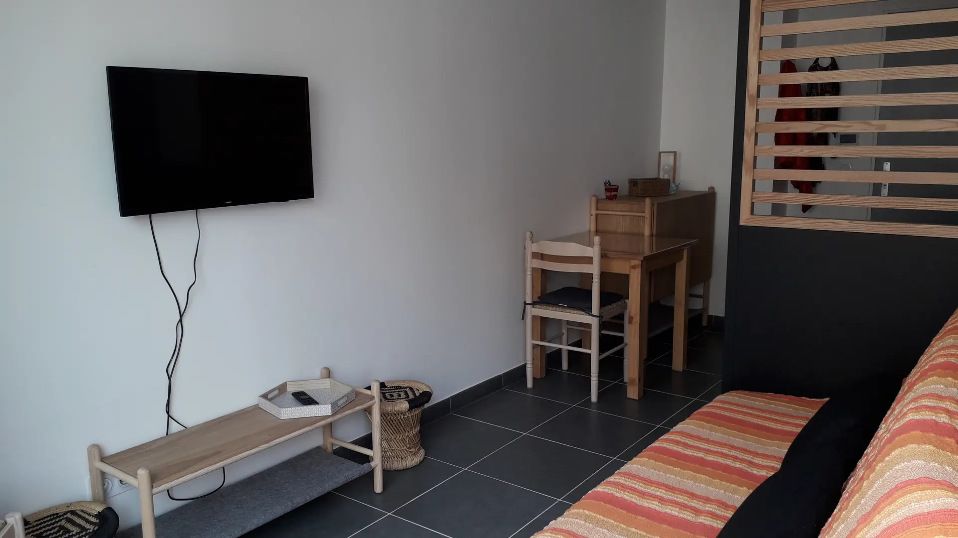 Le salon est équipé d'un canapé en tissu rayé, d'une table basse en bois clair et d'un meuble TV sur lequel est posé un écran plat. Une petite table avec une chaise est également présent.