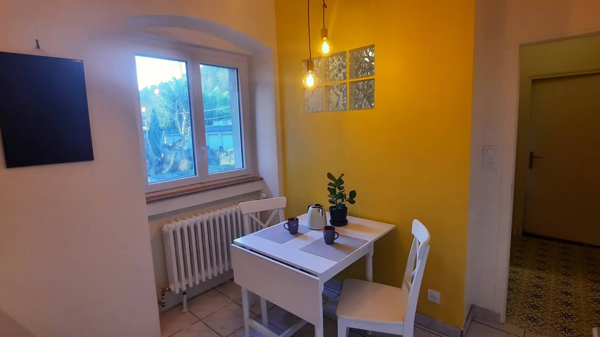 Cette pièce est une cuisine avec un mur jaune de style néo-rétro. Elle est équipée d'une table pouvant accueillir deux personnes, de deux chaises, d'un radiateur, d'une plante et d'un tableau noir. Une fenêtre laisse entrer la lumière naturelle et donne sur l'extérieur.