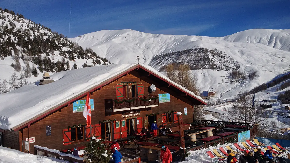 Vue du restaurant, chalet en bois, avec sa terrasse, tables et chaises, chaises lonuges. Montagnes enneigées tout autour et en arrière-plan