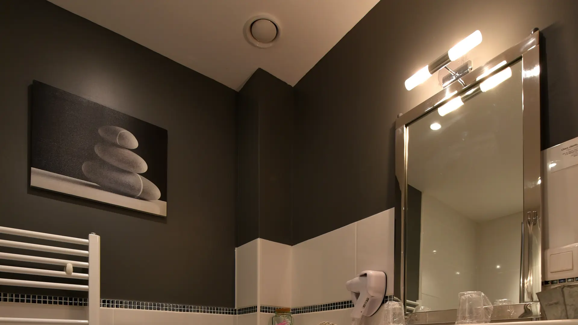 Salle d’eau équipée d’une vasque blanche, d’un sèche serviettes, d’un miroir surmonté d’un néon, de serviettes pliées sous le lavabo, d’un cadre décoratif mural et de toilettes