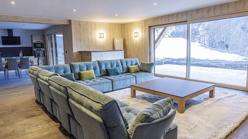 Large, comfortable and bright chalet living room, large corner sofa, rug, coffee table, wooden furniture. Kitchen area with large television and French windows.