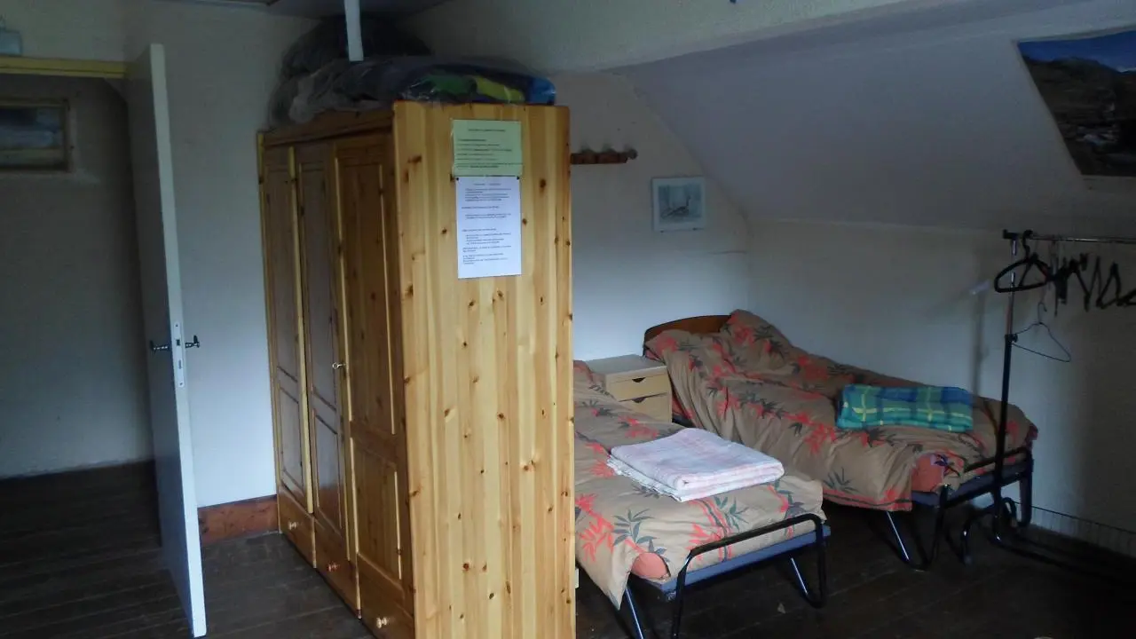 Un dortoir sous combles, on aperçoit deux lits individuels. Une armoire en bois se trouve contre un mur, à proximité de la porte.