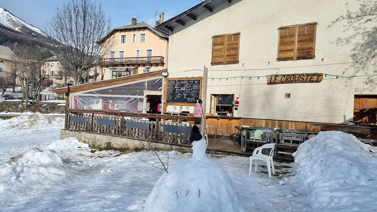 Overview of the restaurant in winter: a two-story stone and wood building with a wooden terrace on the ground floor, tables, and chairs. Snow-covered meadow in front of the restaurant. Wooden sign reading Le Croustet.