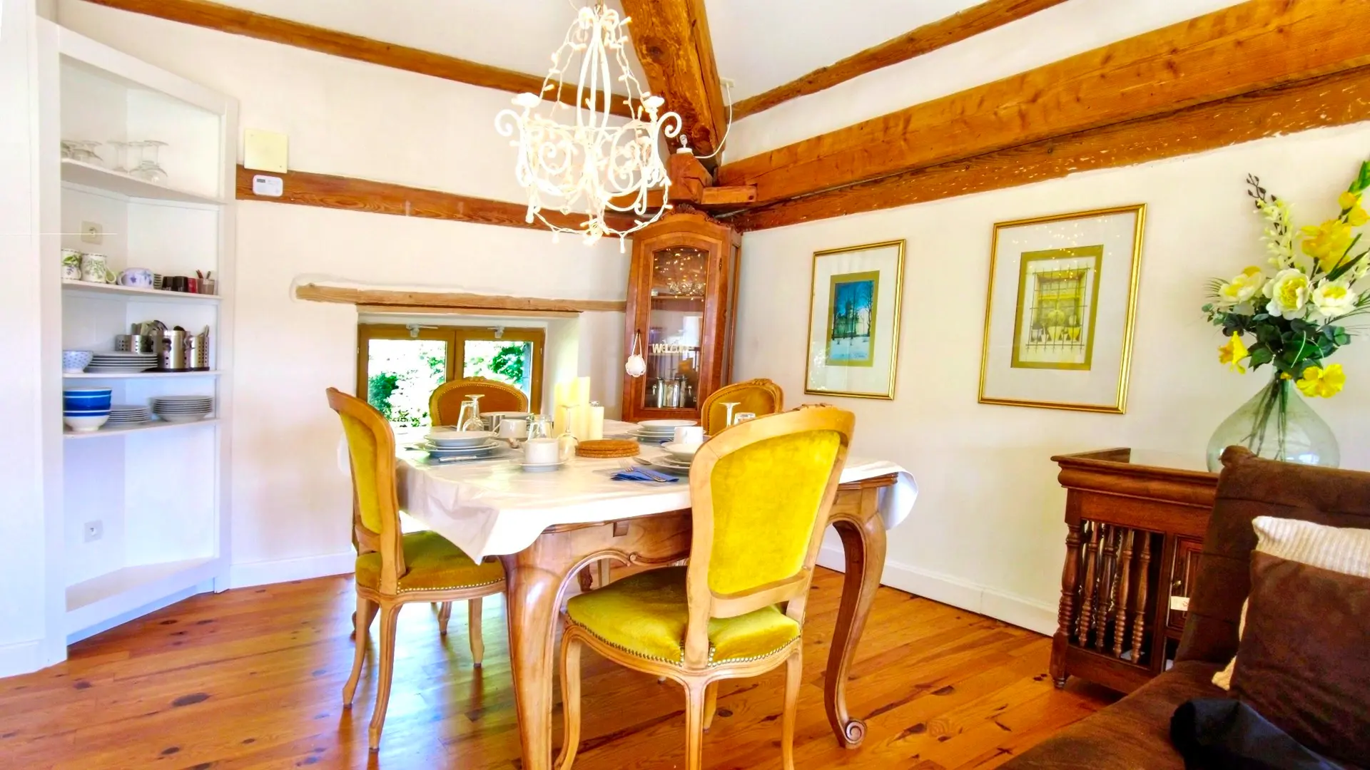 Nous sommes dans la mezzanine dans la partie salle à manger. On y trouve une table dressée pour 4 personnes sur une nappe blanche. Les 4 chaises autour de la table sont en velours jaune. On voit aussi la cuisine américaine, petite mais très fonctionnelle avec tout le nécessaire pour cuisiner. On entrevoit le salon avec la télé encadré d'une bibliothèque. Les poutres de la charpente apparente apporte beaucoup de chaleur sur une décoration vintage. Les poutres principales ont des fanions de couleurs pastels  et un lustre en fer blanc complète la salle à manger