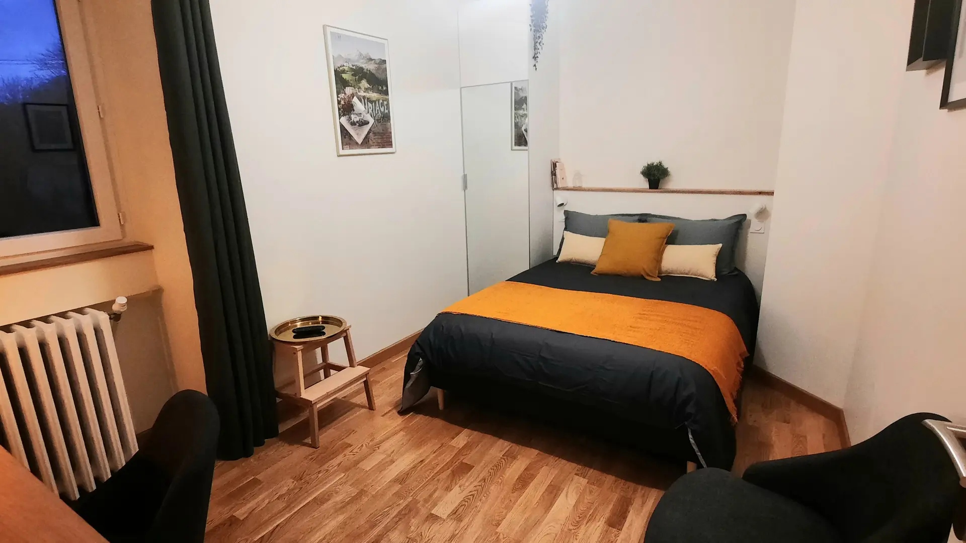 Cette chambre est aménagée avec un lit double habillé d'une couette bleu marine et de coussins jaunes. Deux fauteuils confortables sont placés de part et d'autre d'une petite table basse en bois. Un radiateur est situé sous la fenêtre pour apporter de la chaleur en hiver. Une fenêtre laisse entrer la lumière naturelle et offre une vue sur l'extérieur.