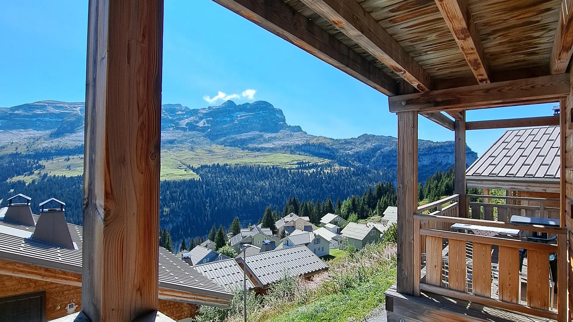 A cozy wooden balcony opens onto a peaceful chalet village nestled against the mountains. It’s a sun-drenched alpine panorama, perfect for a nature getaway.