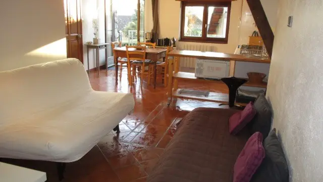 Cette image présente un espace salon, avec un canapé blanc sur la gauche et le coin cuisine avec une table avec des chaises au fond.