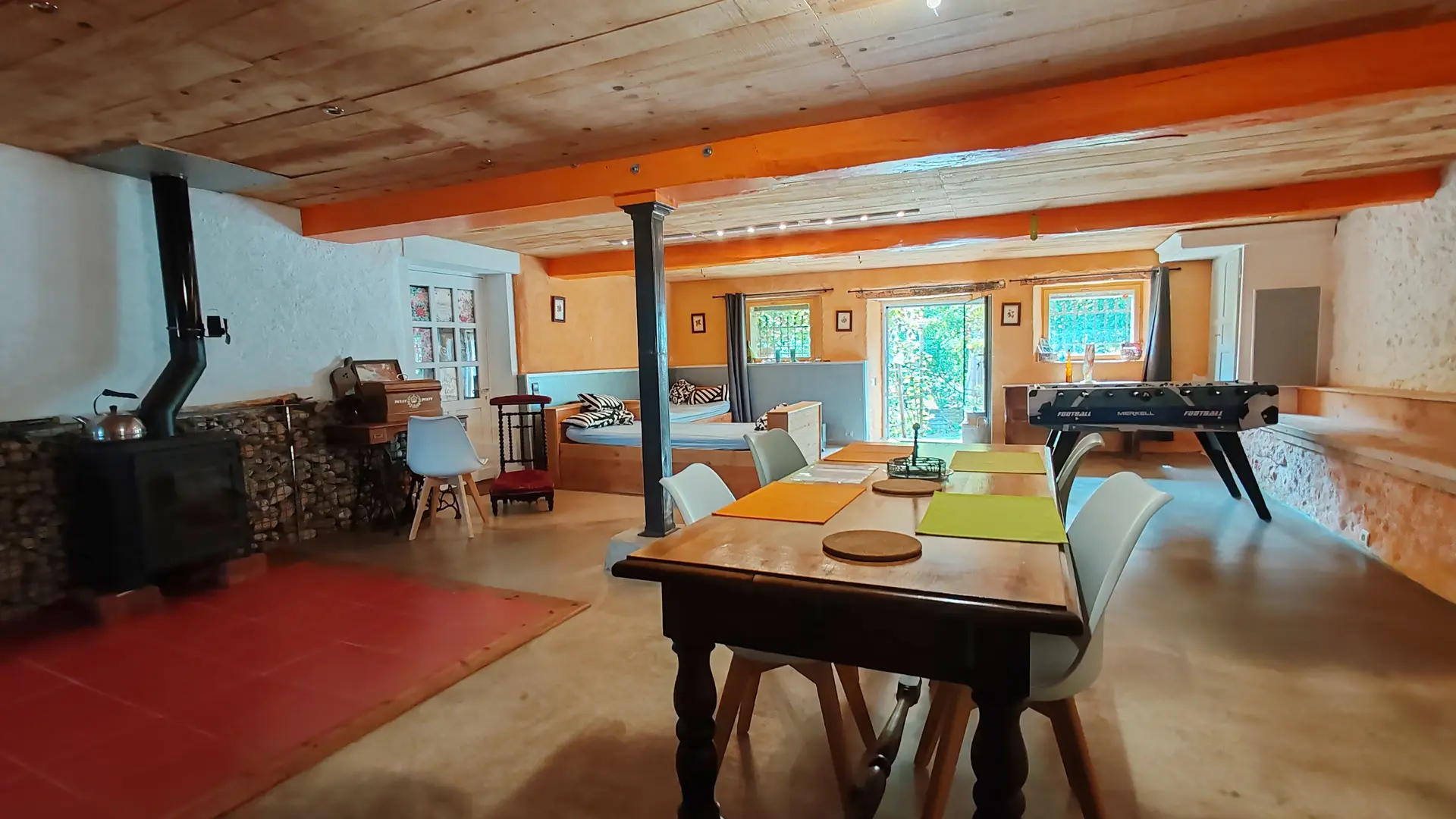 Nous sommes à l'intérieur et une grande pièce de vie se dévoile avec à droite de la porte d'entrée un coin salon et à la gauche un baby-foot. Un coin salle à manger avec sa table permet de prendre ses repas et en face un poèle à bois pour réchauffer les corps et les esprits.