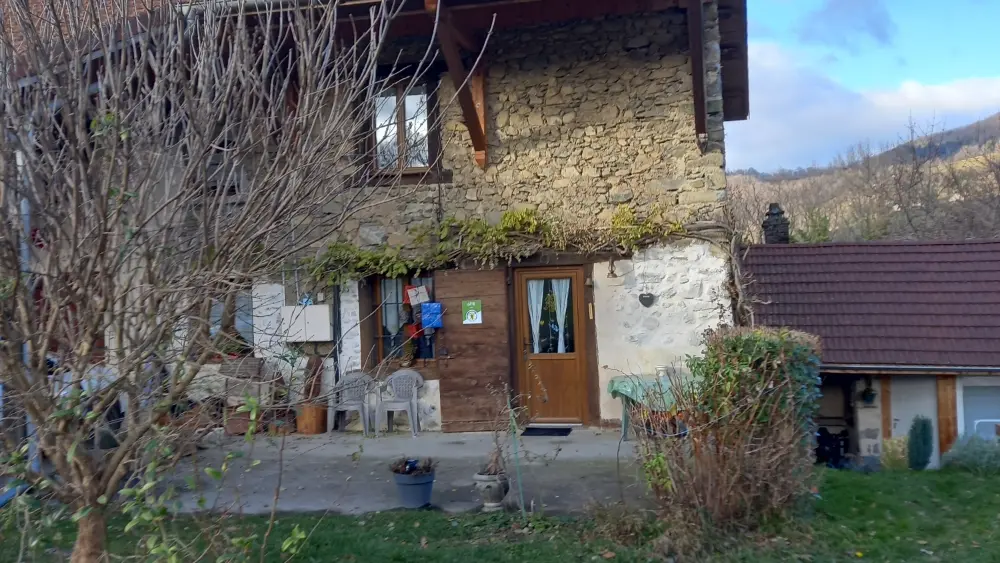 Une grande maison en pierre et bois, avec un toit en pente et un balcon, entourée d'un jardin avec des arbres en premier plan.