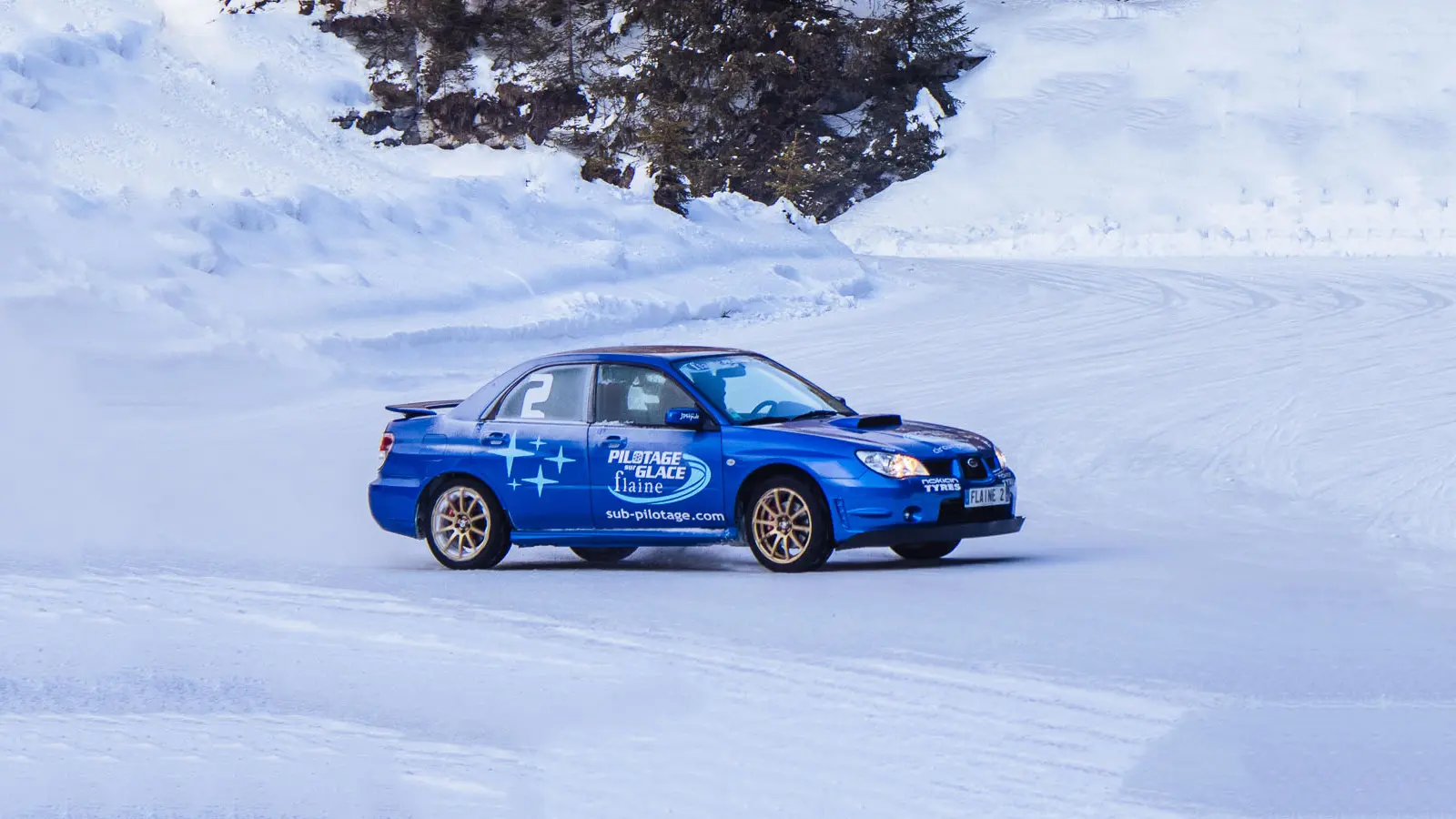 Cette photographie capturent une berline de sport bleue lancée à vive allure sur le circuit de glace de Flaine, entouré de sapins et de parois rocheuses enneigées.