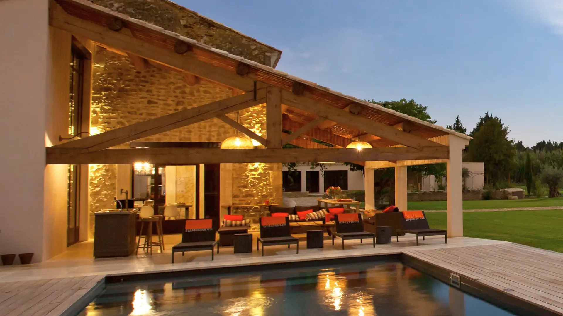 Piscine éclairée le soir, terrasse couverte en pierre et bois, ambiance conviviale.