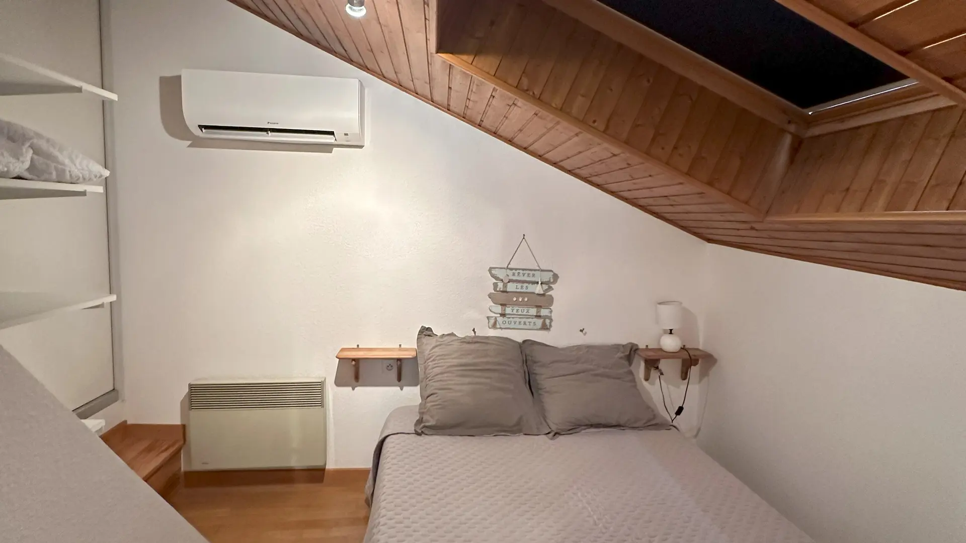 La pièce est une chambre sous les combles avec un lit double placé au centre. Un velux occupe une grande partie du plafond, laissant entrer une lumière naturelle abondante. Le sol est en parquet.