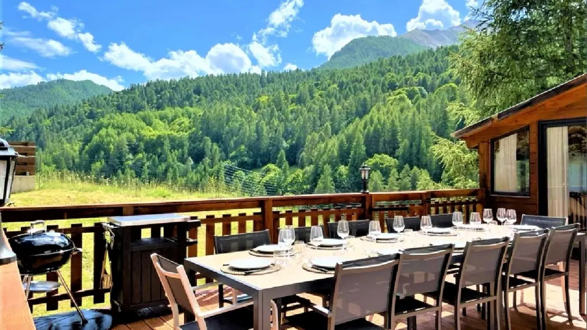 Terrasse en bois du chalet avec grande table d'extérieur, transat, véranda, vue dégagée avec paysage d'été