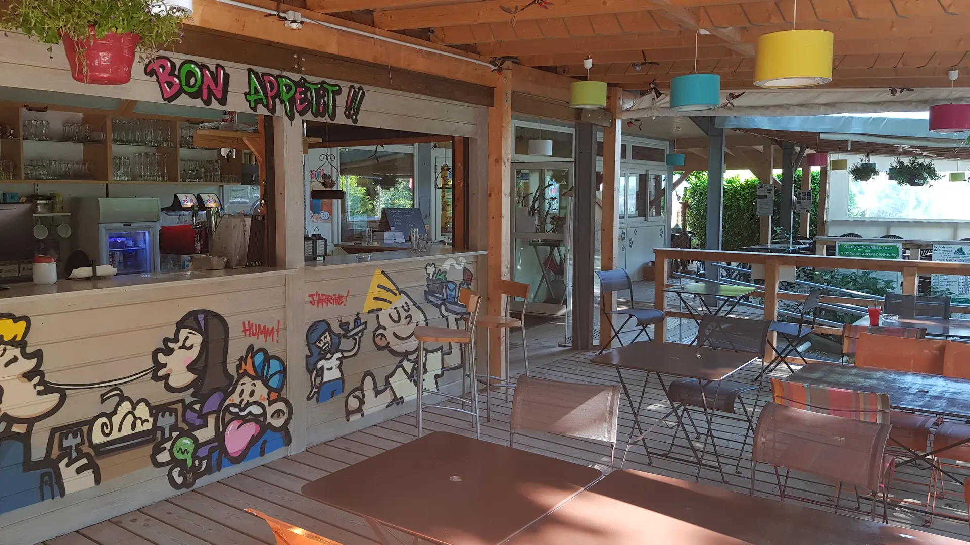 Un espace de restauration en plein air, aménagé sous une structure en bois avec une terrasse, comprenant un bar et des tables avec chaises. Des décorations murales colorées et ludiques animent l'espace.