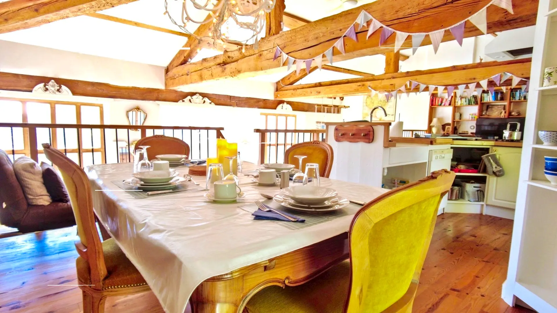 Nous sommes dans la mezzanine dans la partie salle à manger. On y trouve une table dressée pour 4 personnes sur une nappe blanche. Les 4 chaises autour de la table sont en velours jaune. On voit aussi la cuisine américaine, petite mais très fonctionnelle avec tout le nécessaire pour cuisiner. On entrevoit le salon avec la télé encadré d'une bibliothèque. Les poutres de la charpente apparente apporte beaucoup de chaleur sur une décoration vintage. Les poutres principales ont des fanions de couleurs pastels  et un lustre en fer blanc complète la salle à manger