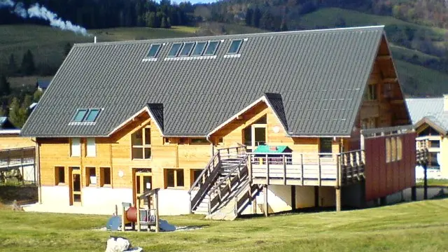 Bâtiment en bois avec le toit en tôles grises, pourvu de fenêtres de toit, un escalier et une terrasse en bois sur la droite, une petite aire de jeux extérieure devant le bâtiment