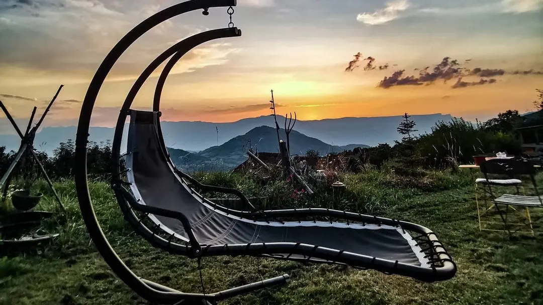 Une chaise suspendue en forme de croissant de lune est placée sur une pelouse verdoyante, face à un paysage montagneux au coucher du soleil. Le ciel est parsemé de nuages colorés, et l'on distingue au loin des arbres et d'autres éléments de mobilier de jardin.