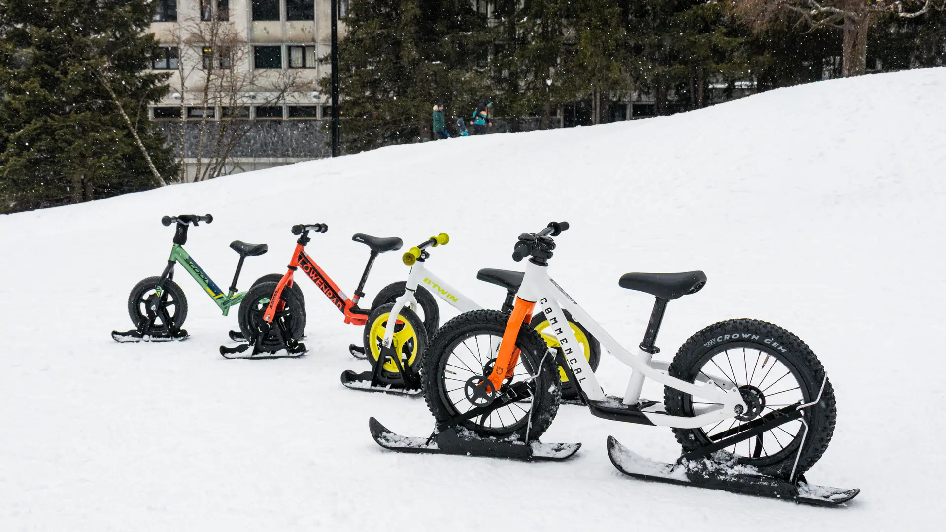 Quatre vélos-ski de différentes couleurs (vert, rouge, blanc et jaune) sont alignés dans la neige sur une pente douce.