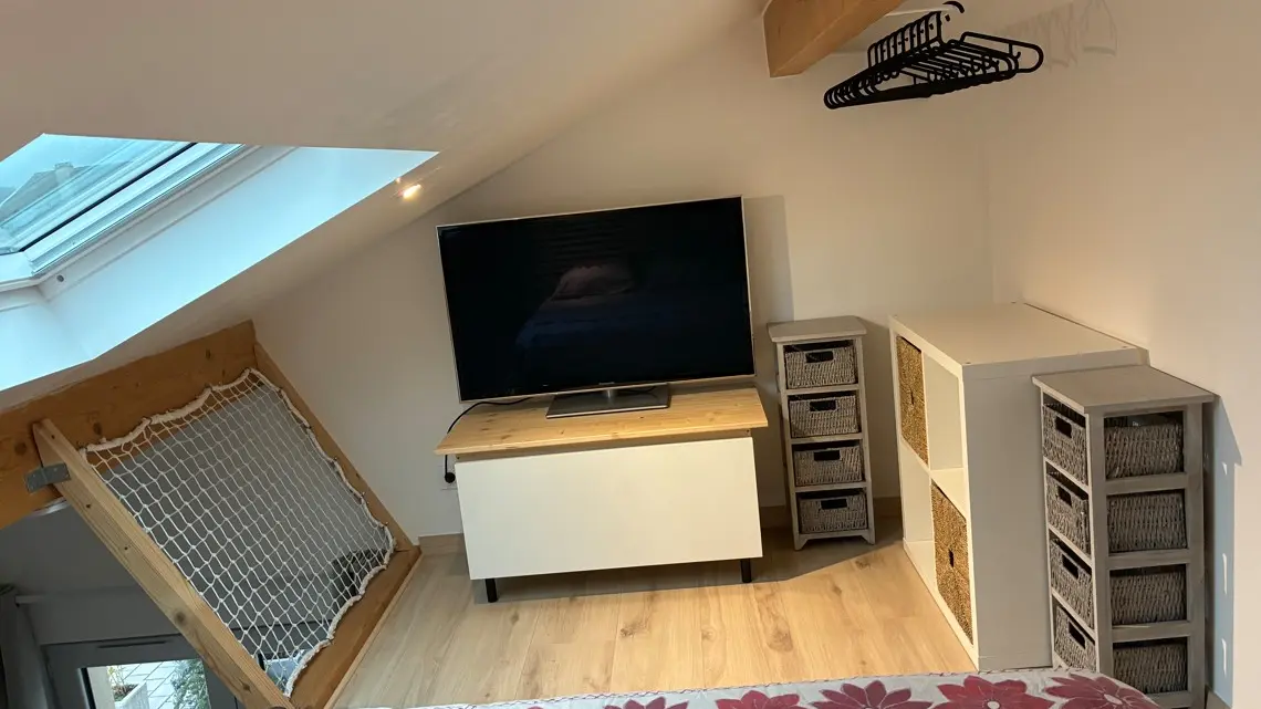 Une chambre mansardée avec un téléviseur posé sur un meuble blanc, des rangements à cases sur la droite et une fenêtre de toit apportant de la lumière sur un parquet clair.