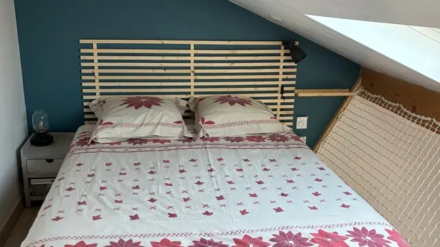 Un lit double avec une couette blanche à motifs de fleurs roses est placé sous une pente de toit, contre un mur bleu canard orné d'une tête de lit en bois clair.