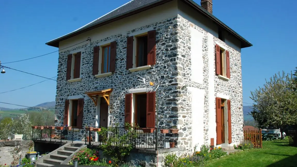 C'est une maison de deux étages, construite en pierres apparentes de couleur claire, avec un toit à quatre pans. Elle possède un jardin avec une pelouse et des fleurs, ainsi qu'une terrasse accessible par quelques marches.