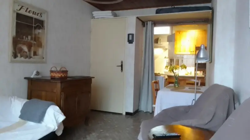 Un coin salon avec des fauteuils et un canapé, faisant face à une commode en bois et une porte beige, tandis qu'une niche ouverte à droite révèle une petite cuisine et une table à manger.