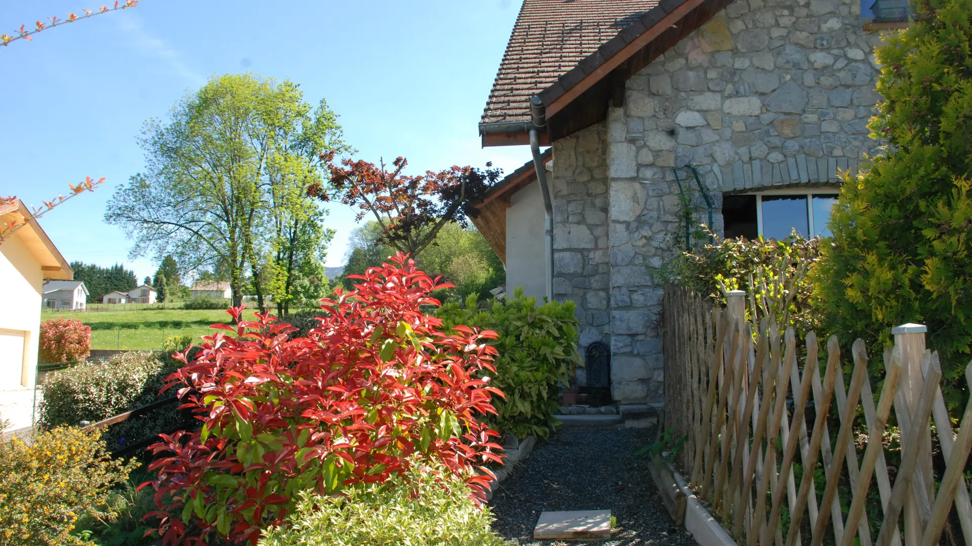 Cette habitation est caractérisée par ses murs en pierre et son toit en pente. Un jardin agrémente la façade, avec un chemin gravillonné menant à l'entrée. Des arbustes, notamment un arbuste aux feuilles rouge vif, bordent le chemin.