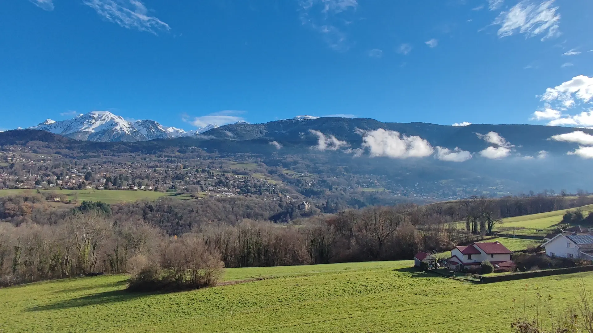Une vue panoramique de la chaîne de Belledonne montre des sommets enneigés et la station de Chamrousse dominant des collines verdoyantes sous un ciel bleu.