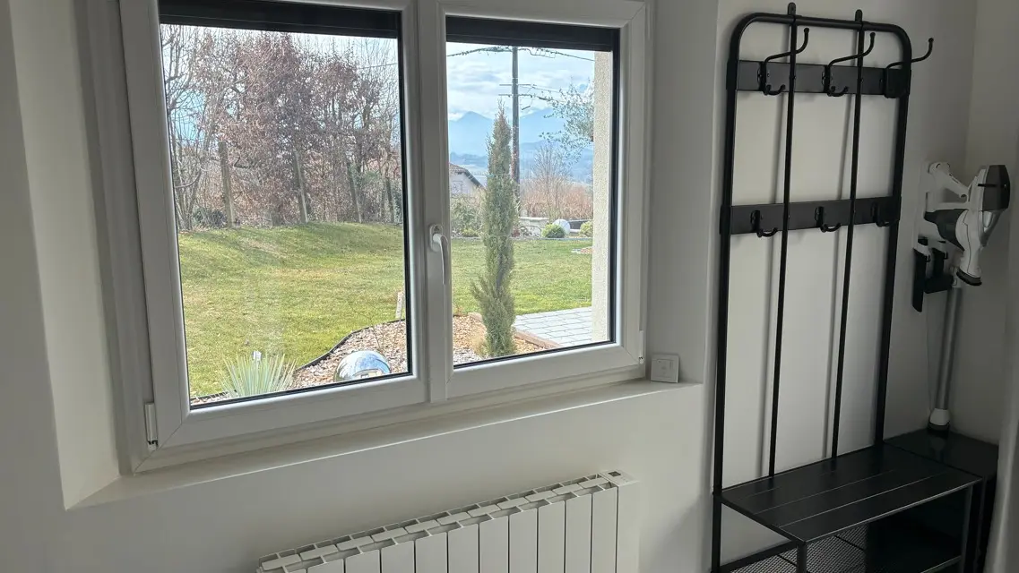 L'entrée de la maison dispose d'un vestiaire mural en métal noir avec banc intégré, situé à côté d'une fenêtre blanche offrant une vue dégagée sur le jardin et d'un radiateur blanc.