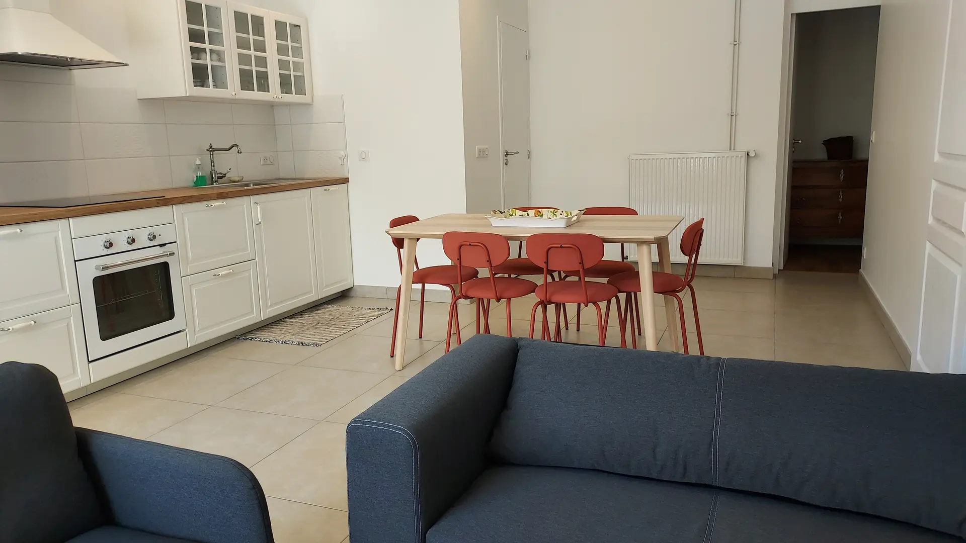a pièce est un espace ouvert moderne combinant une cuisine claire avec des armoires blanches, un coin repas doté d'une table en bois et de chaises rouges, et un salon avec des canapés sombres en premier plan.