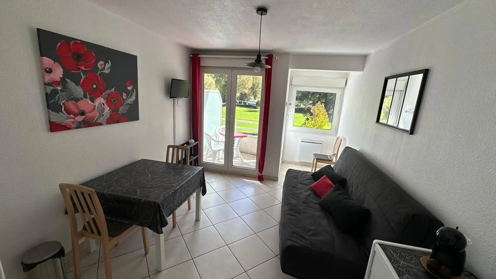 La pièce est un salon meublé avec un canapé-lit et un espace repas ou bureau, orné de deux tableaux, et donnant sur l'extérieur par une porte-fenêtre centrale.