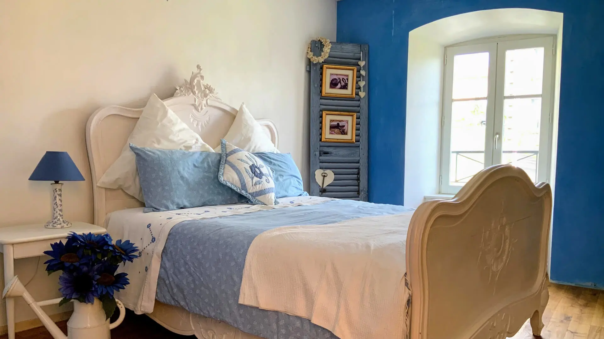 C'est la chambre bleue avec son lit vintage restauré en blanc. Un pan de mur près de la fenêtre est en bleu azur, l'encadrement de fenêtre reste en blanc pour accrocher la lumière et la restituer à la pièce. Le reste des murs est en blanc cassé. Le sol est un plancher de couleur miel. Le linge de lit est assorti à la couleur de la chambre. Les éléments de décoration sont peu nombreux. Sur le côté droit du lit, un volet bleue est revisité en élément de décoration où sont accrochés des tableaux et autres éléments de décoration. Sur le côté gauche du lit, une table de nuit de la même couleur que le lit accueille une lampe de chevet bleue et au pied de la table de chevet, un arrosoir peint en blanc cassé est détourné en contenant de fleurs bleues. Cette ambiance cosy se retrouve dans les deux autres chambres tant au niveau des couleurs, de l'harmonisation des éléments de décorations et linge de lit.