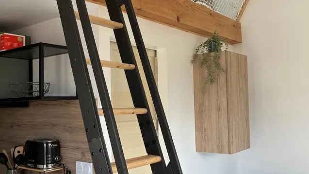 Une échelle de meunier en bois bicolore, noir et naturel, mène à une mezzanine sécurisée par un filet et des poutres apparentes, surplombant une pièce de vie avec un canapé gris.