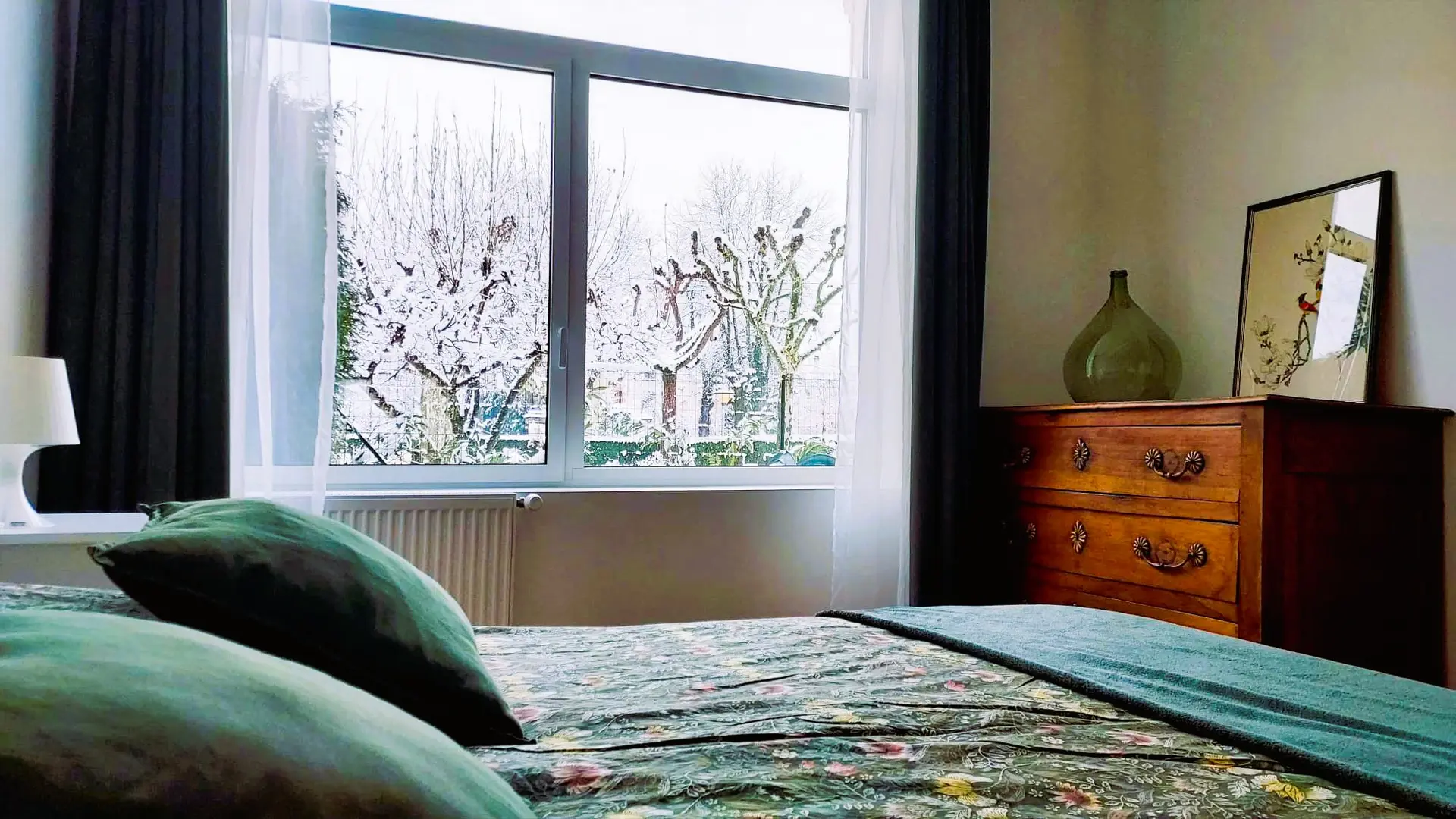 L'intérieur d'une chambre à coucher avec un lit doté de coussins verts et d'une couverture turquoise, faisant face à une grande fenêtre qui donne sur un paysage enneigé, et un meuble en bois clair avec un vase vert est visible à droite.