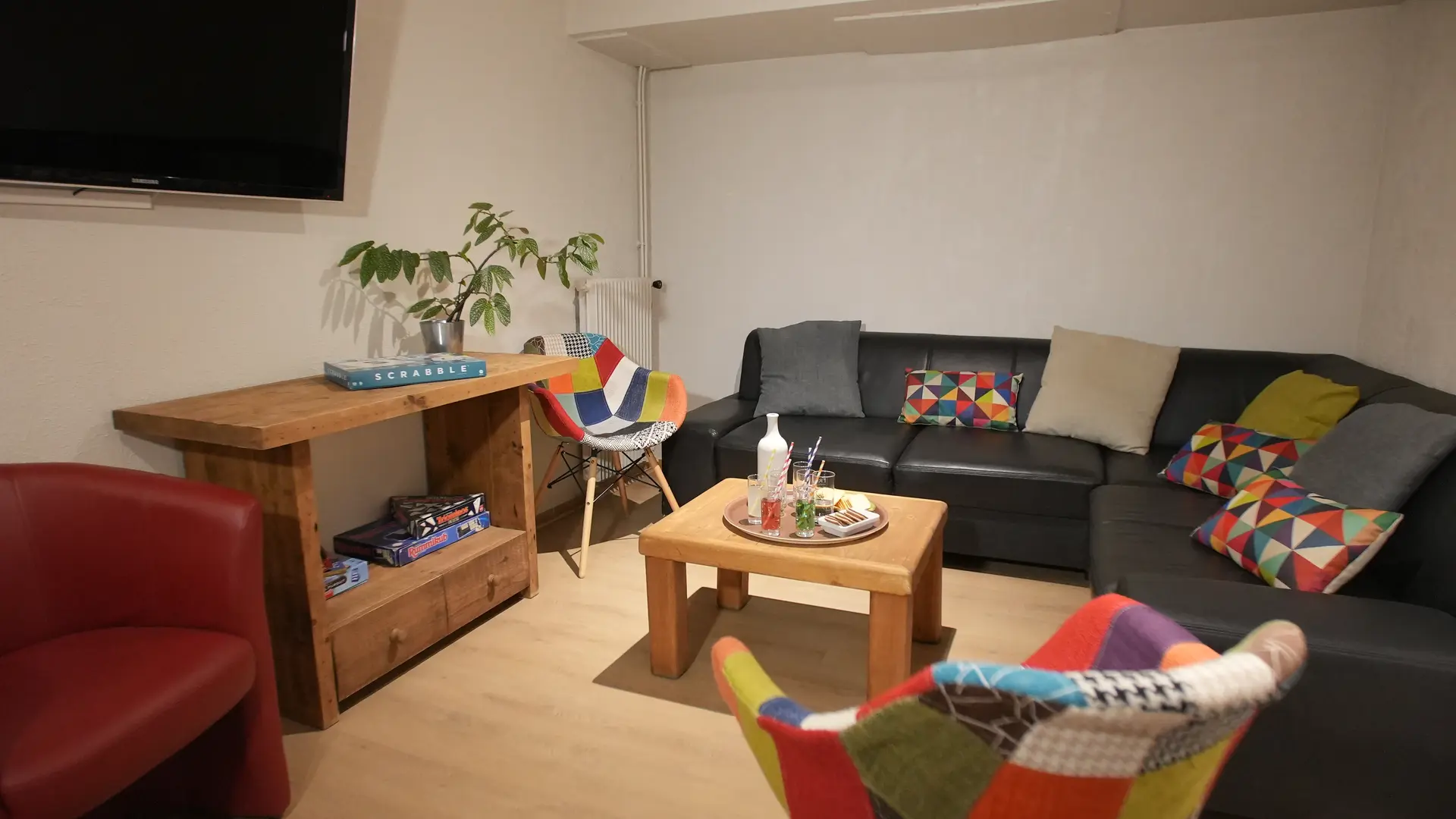 Canapé d’angle avec coussins colorés, deux chaises patchwork et table centrale en bois surmontée d’un plateau avec verres et pailles. Une plante verte et un écran de télévision à gauche complètent l’aménagement.