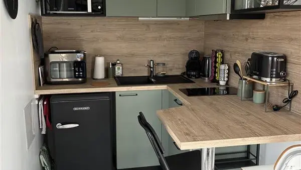 Une cuisine équipée en angle avec des meubles vert sauge et un plan de travail en bois clair comprend un petit réfrigérateur noir, un évier encastré et un retour de comptoir servant de table avec deux chaises hautes.