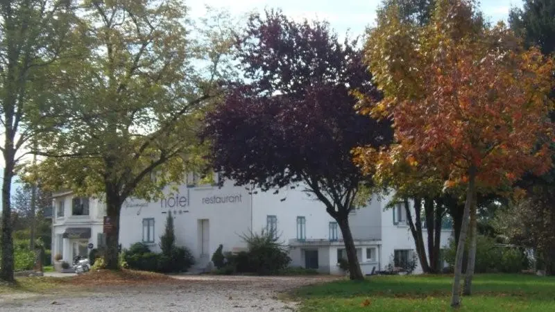 L'hôtel est situé dans un environnement verdoyant. De grands arbres entourent le bâtiment, offrant de l'ombre et créant une atmosphère paisible. Les feuilles des arbres sont de différentes couleurs, ce qui suggère que la photo a été prise à l'automne. Un chemin mène à l'hôtel, traversant une pelouse verdoyante.