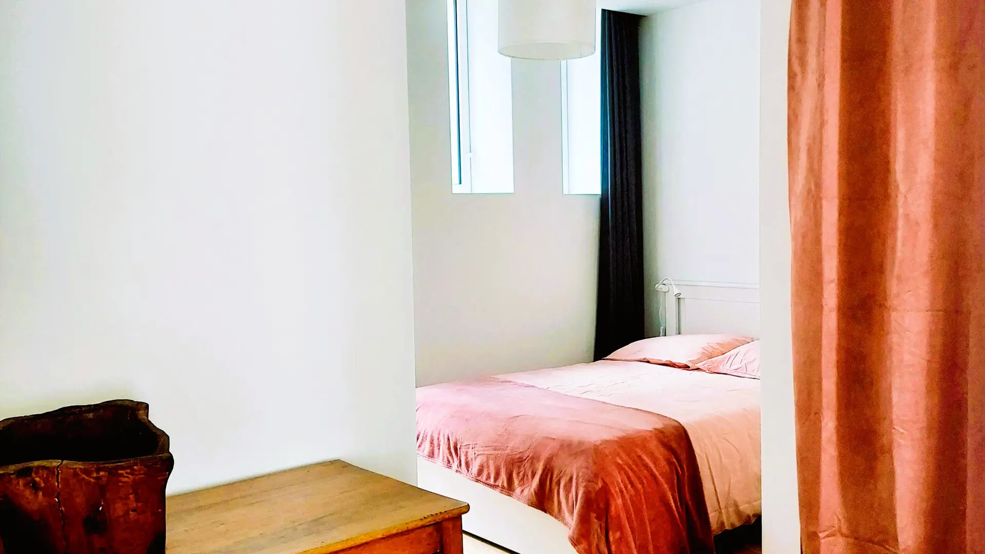 Un intérieur lumineux avec un plancher en bois, mettant en avant une commode en bois foncé au premier plan et, à l'arrière-plan, une chambre partiellement visible derrière un rideau orange, où l'on distingue un lit avec une literie rose et orange.