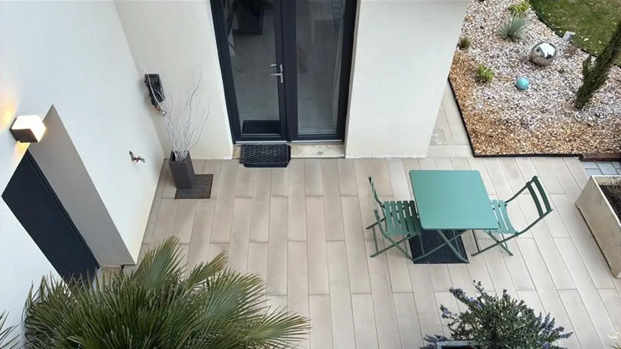 Une terrasse en dalles claires accueille un petit salon de jardin en métal vert composé d'une table et de chaises, située devant une porte vitrée à cadre noir et bordée par un massif de graviers décoratifs.