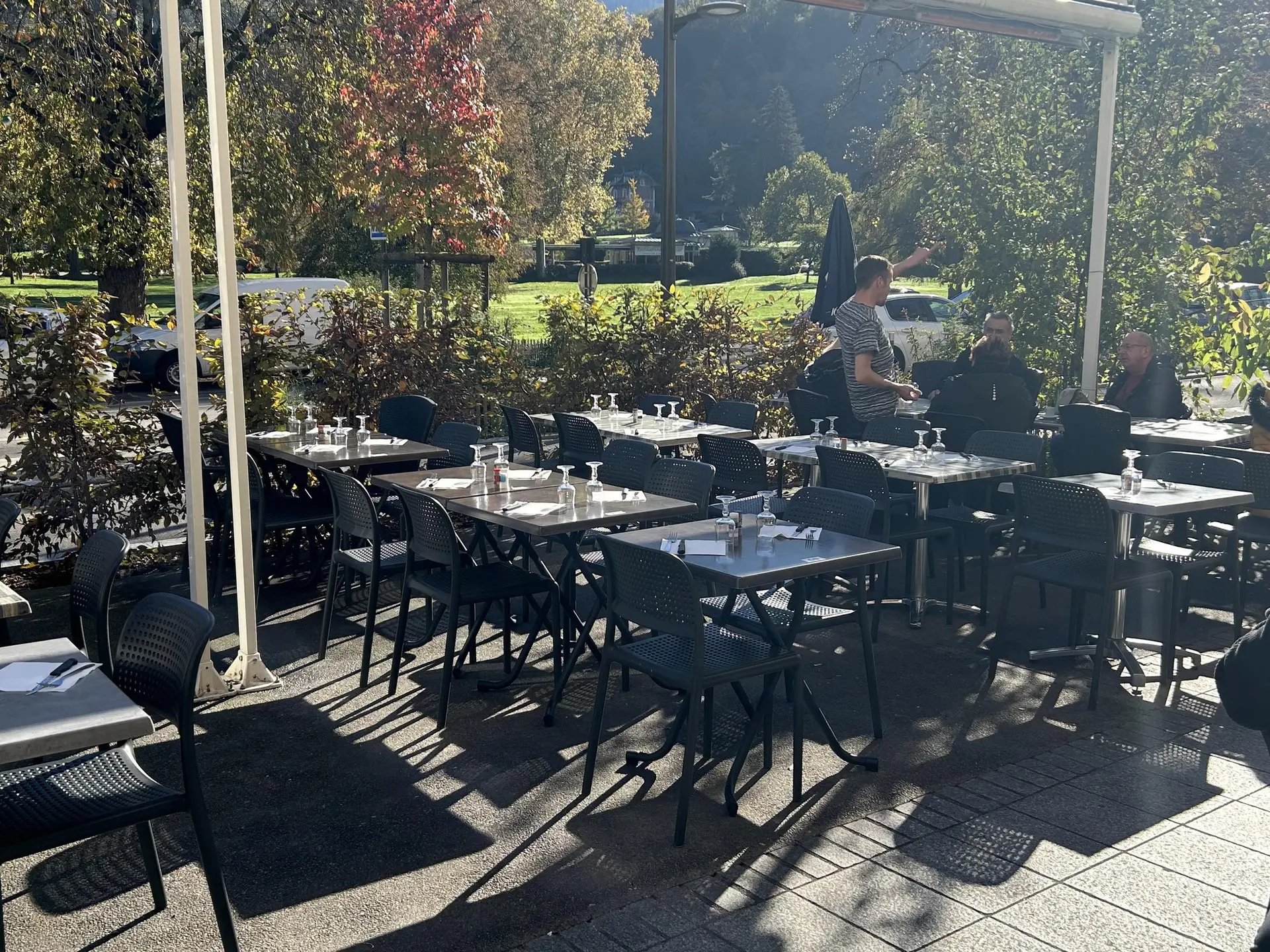 Terrasse de café/restaurant ensoleillée avec une vue sur un parc avec des grands arbres.