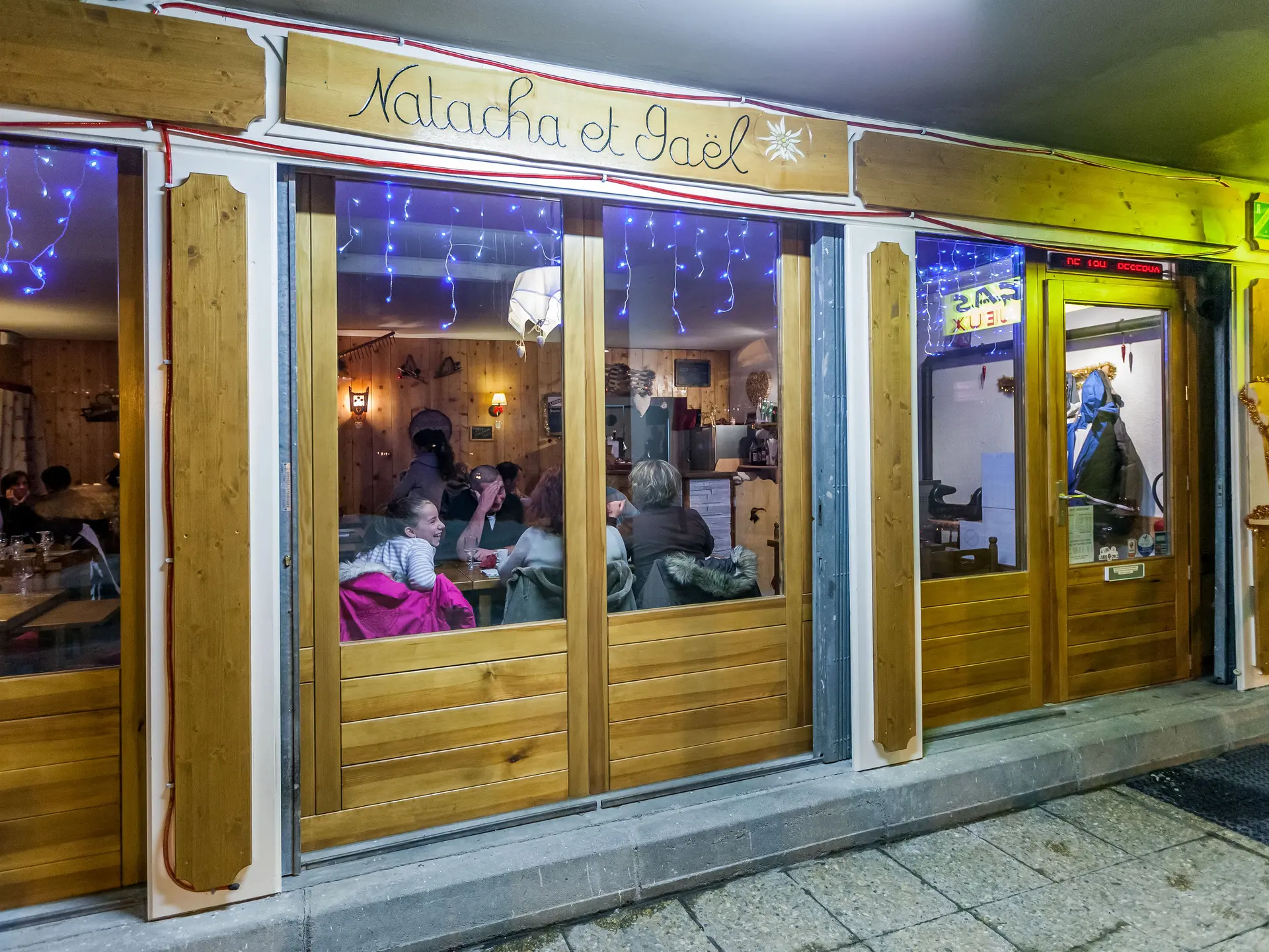 Vitrine du restaurant, façade en bois avec inscription gravée Natacha et Gaël. A l'intérieure table et chaises, ambiance confortable