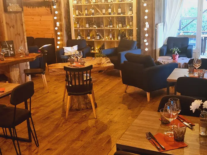 Intérieur du restaurant, ambiance chaleureuse avec fauteuils confortables, intérieur bois, tables en bois, bouteilles de vin en présentoir, porte-fenêtre donnant sur la terrasse de l'établissement