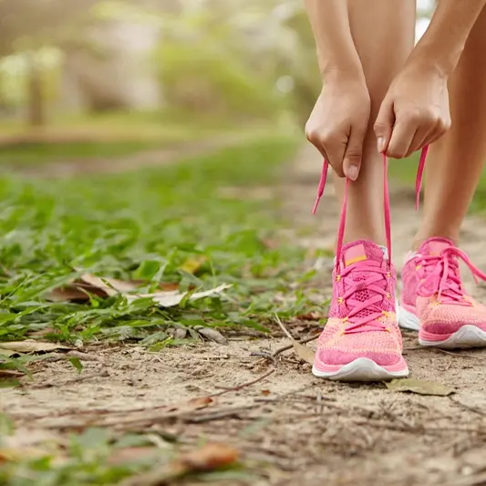 Caucasian athletic woman tying laces on her pink running shoes before jogging standing on footpath in forest. Female runner lacing her sneakers while doing workout in rural area. Film effect